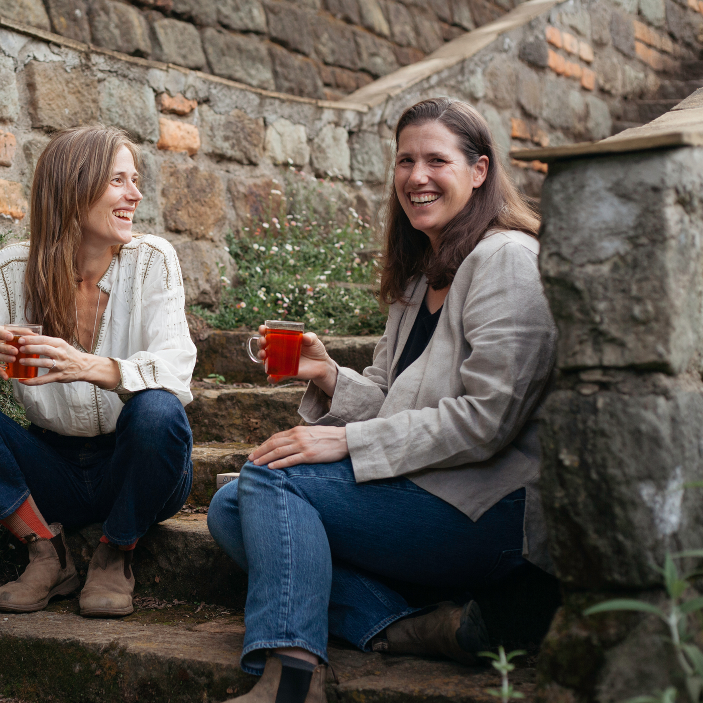 Ajiri Tea co-founders Kate and Sara laughing on stone steps in Kenya, each holding a glass of tea