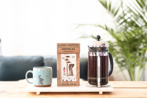 Box of Ajiri Kenyan AA whole bean coffee between a blue elephant mug and a French press of coffee