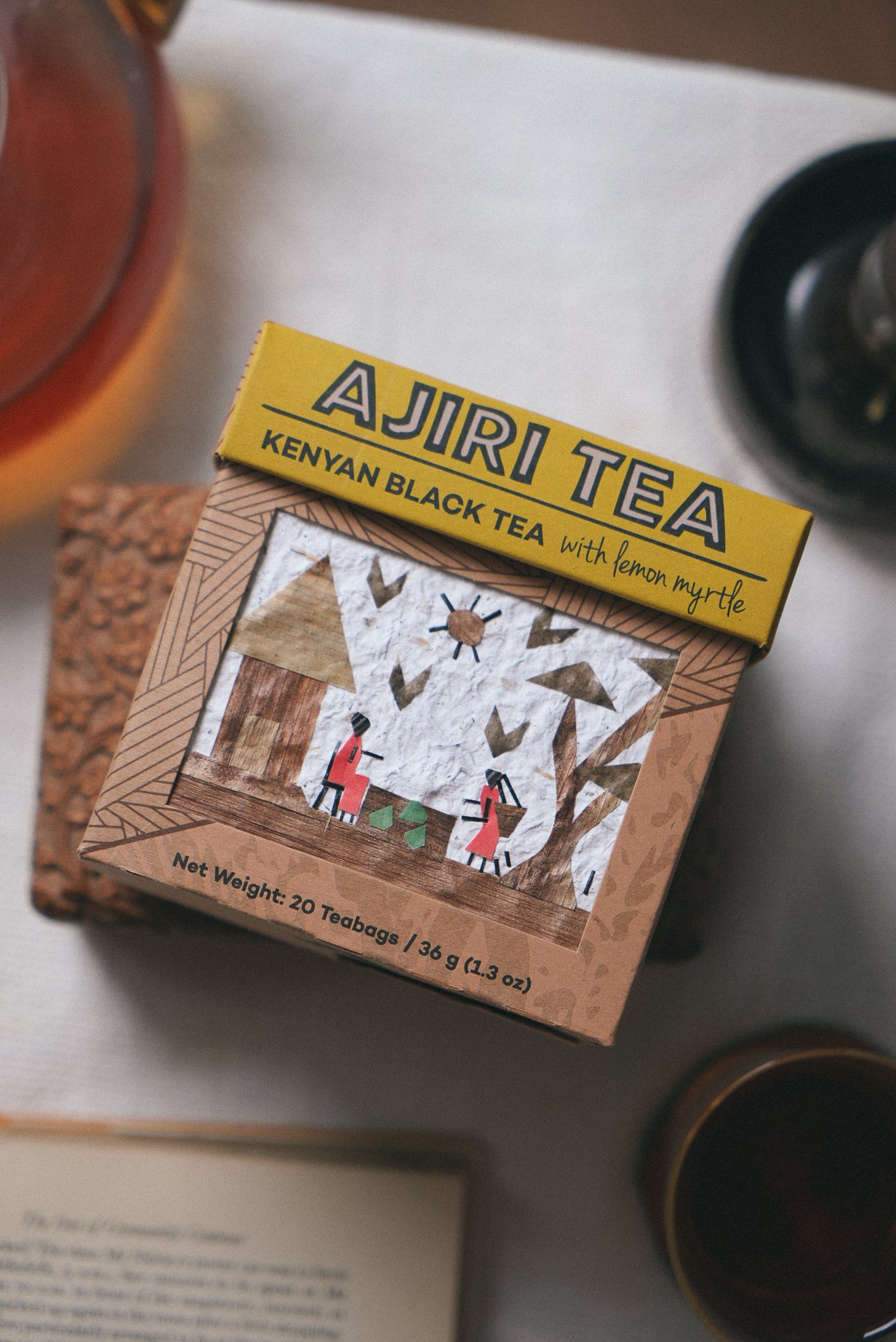 A box of Ajiri Kenyan Black Tea with lemon myrtle, with its handmade art panel, beside a glass teapot and cups