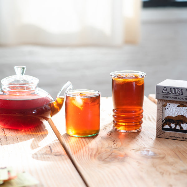 Glass teapot and two glasses of Kenyan black tea beside a box of Ajiri Kenyan Black Tea on a sunlit wooden table