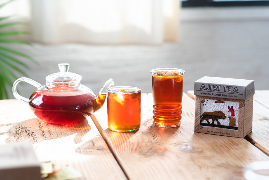 Glass teapot and two glasses of Kenyan black tea beside a box of Ajiri Kenyan Black Tea on a sunlit wooden table